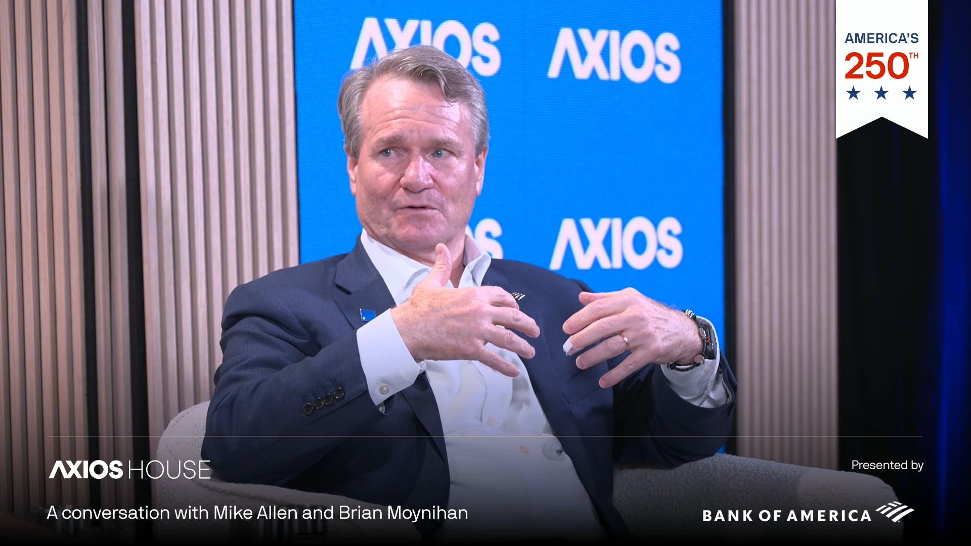 Man in a dark suit gesturing while speaking on a panel. Blue Axios backdrop with the Axios logo; bottom overlay reads "A conversation with Mike Allen and Brian Moynihan" and shows Bank of America branding.