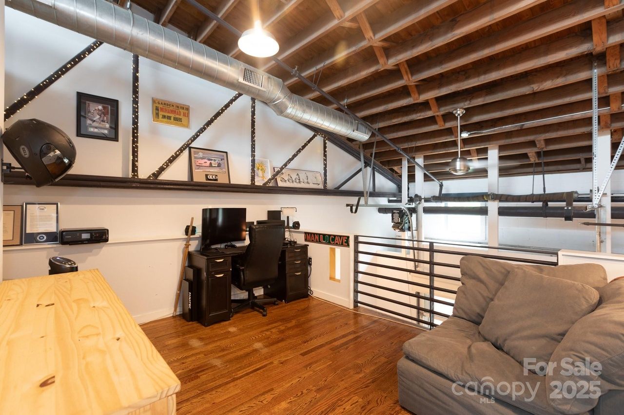 Loft space with exposed wooden beams and metal pipes, featuring a black desk and chair with a computer, a gray couch, wooden floor, and framed pictures on a white wall.