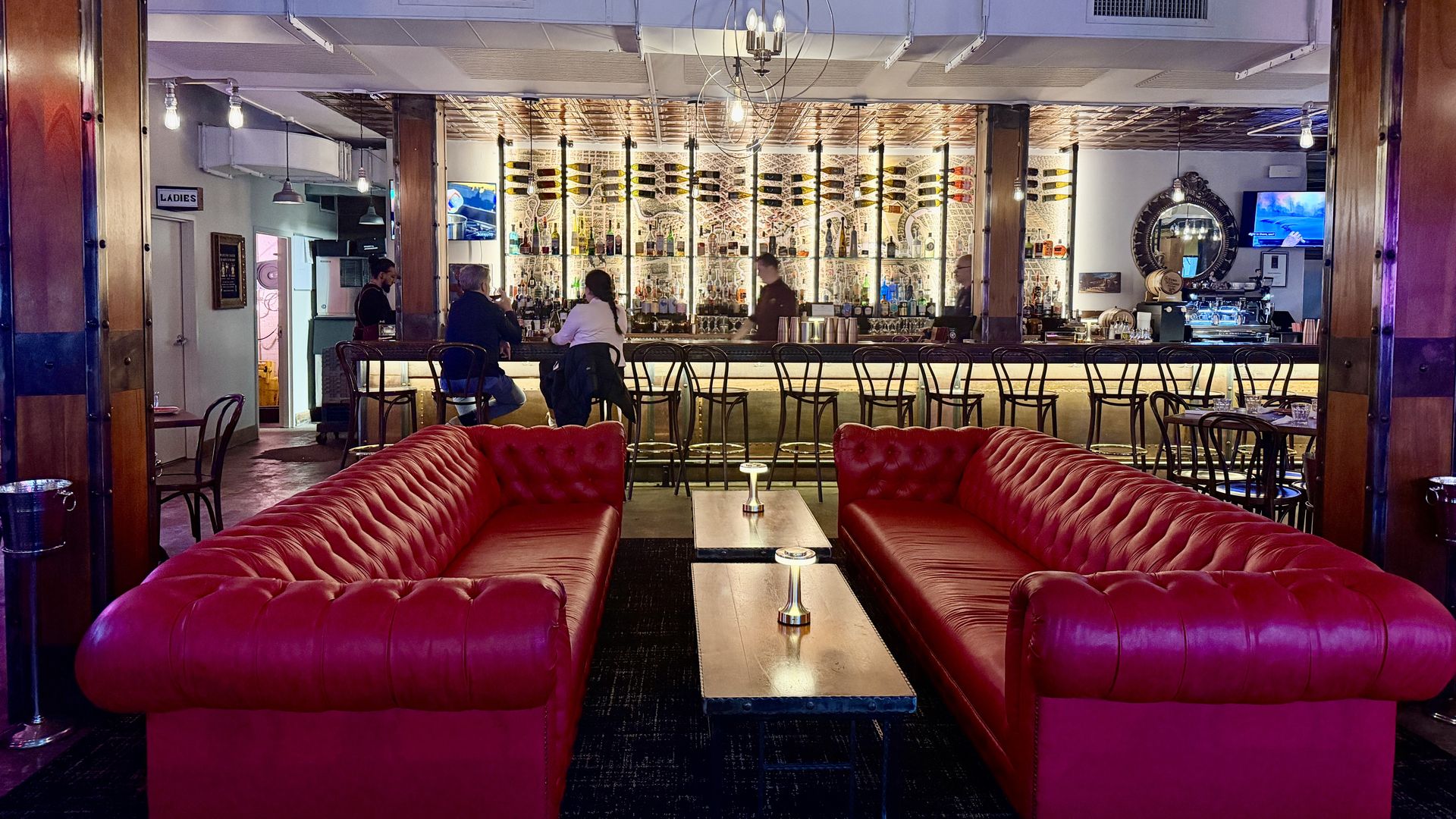 Interior of a cozy bar with two red leather sofas facing wooden tables, people sitting at the bar with bottles and warm lighting behind, and wooden columns and ceiling beams.
