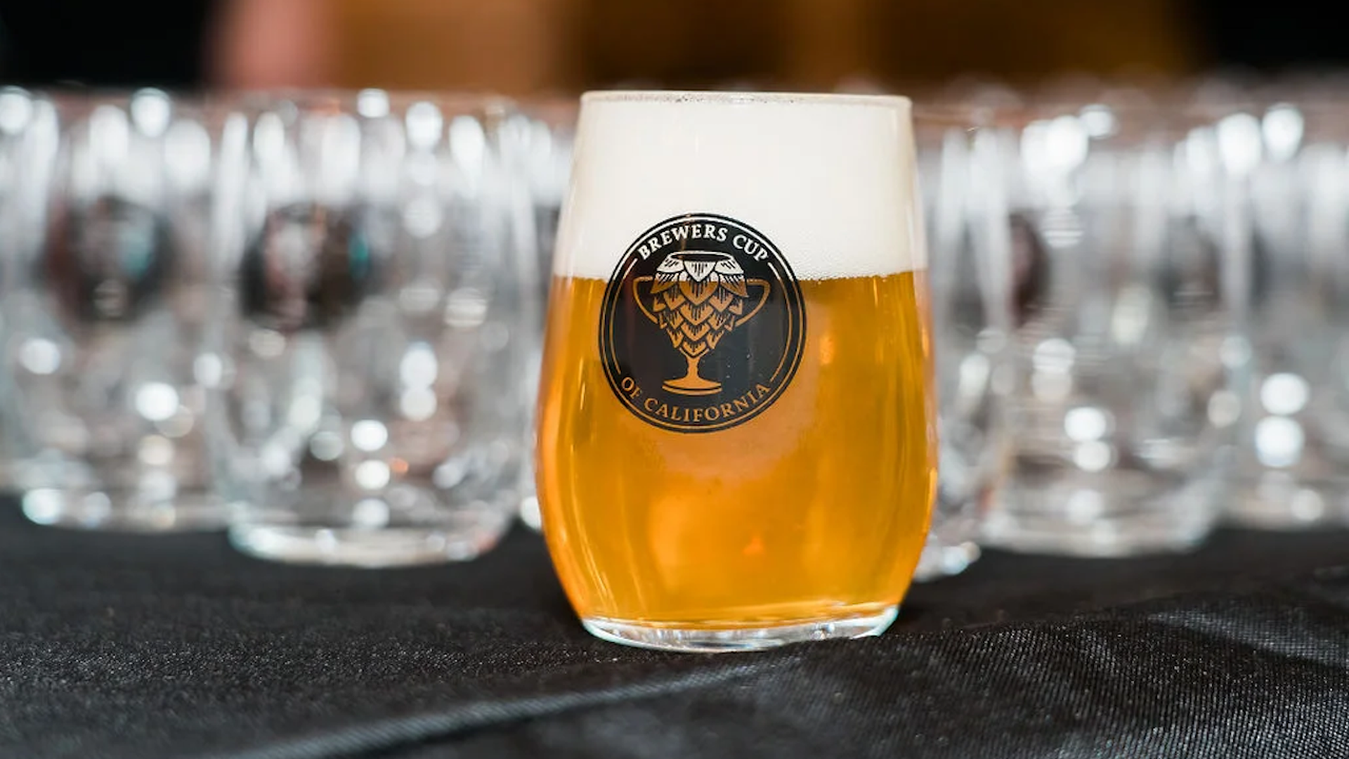 A sample cup of beer with a Brewers Cup of Claifornia logo and empty versions of the glass behind it