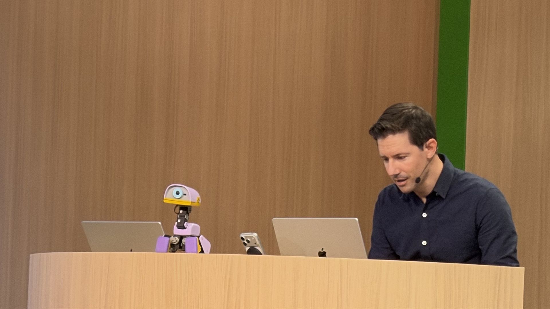 Man in a dark shirt sits at a wooden desk with two laptops; a small pink robot sits on the desk. Bright orange and green panels behind him, with exposed beams and a large window in the background.