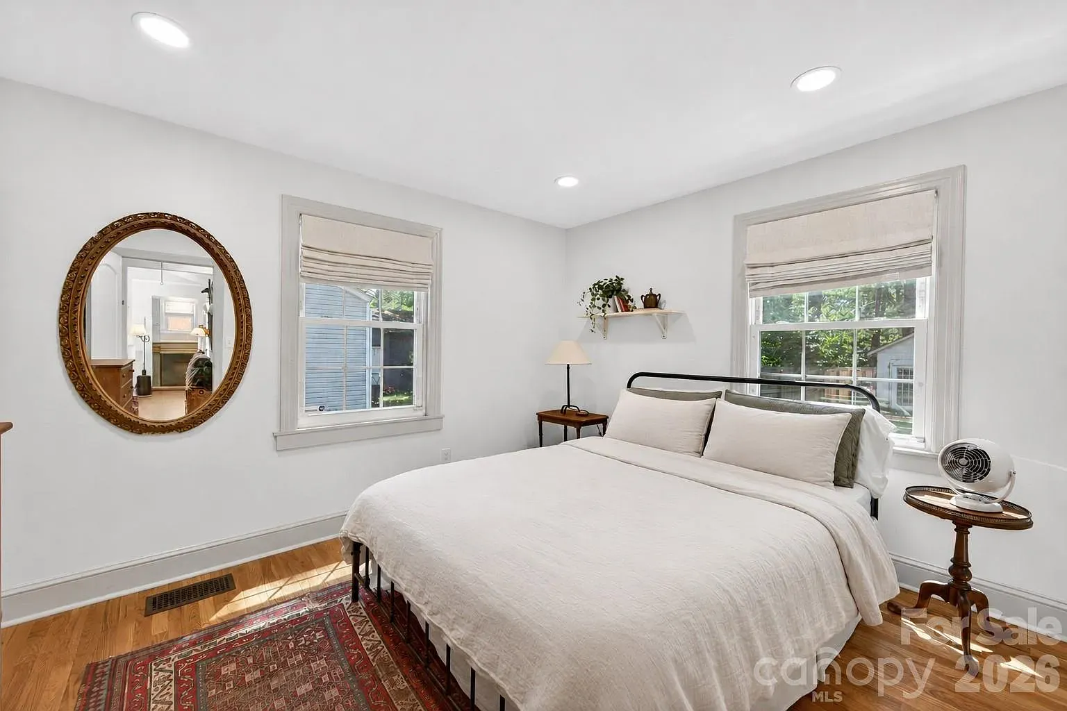 houses for sale in charlotte. Bright bedroom with white bedding on a black metal bed frame, two windows with beige Roman shades, a round ornate wooden mirror on the left, a red patterned rug, and small side tables with a lamp and a fan.