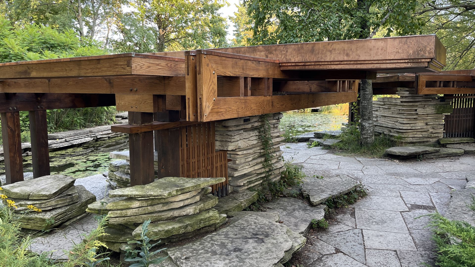 Wooden bridge structure with thick beams and stacked stone supports over a pond with lily pads, surrounded by green trees and stone pathways in a garden setting.