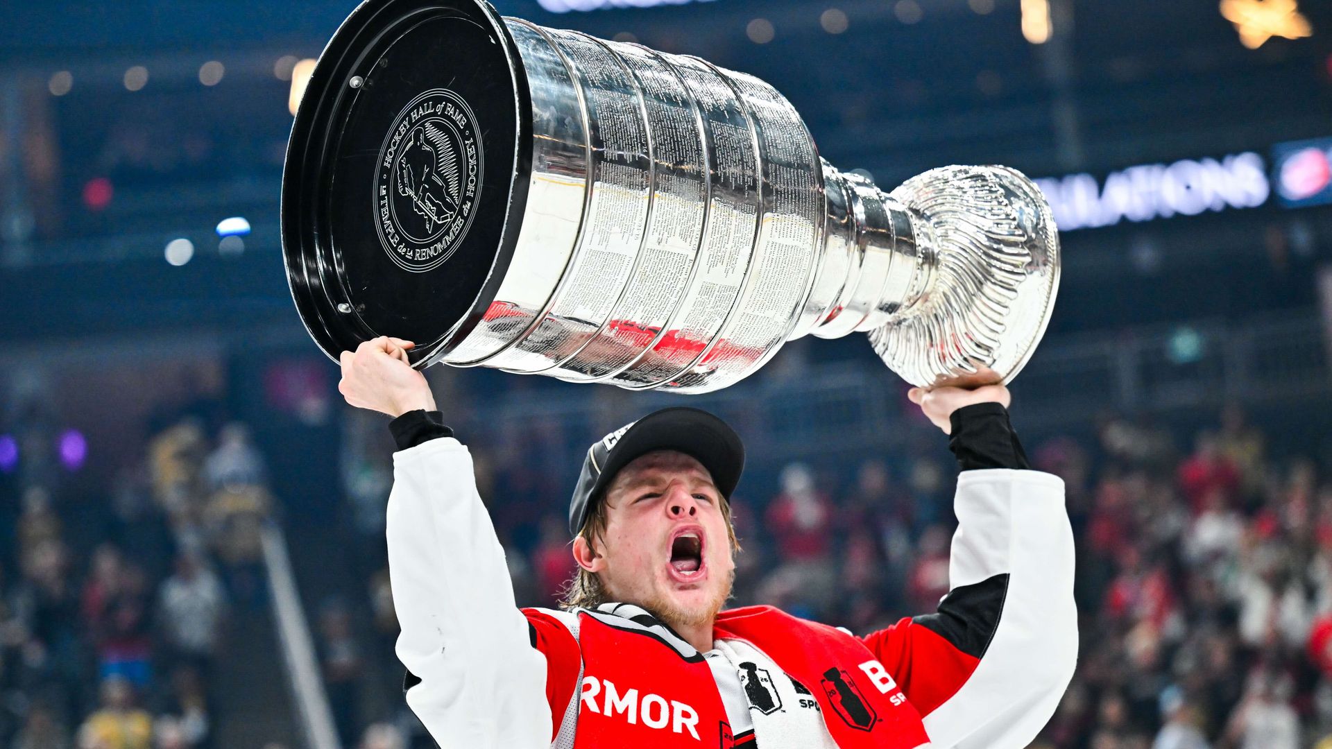 A hockey player in a red and white jersey lifts a large, gleaming Stanley Cup above their head in a crowded arena, celebrating.