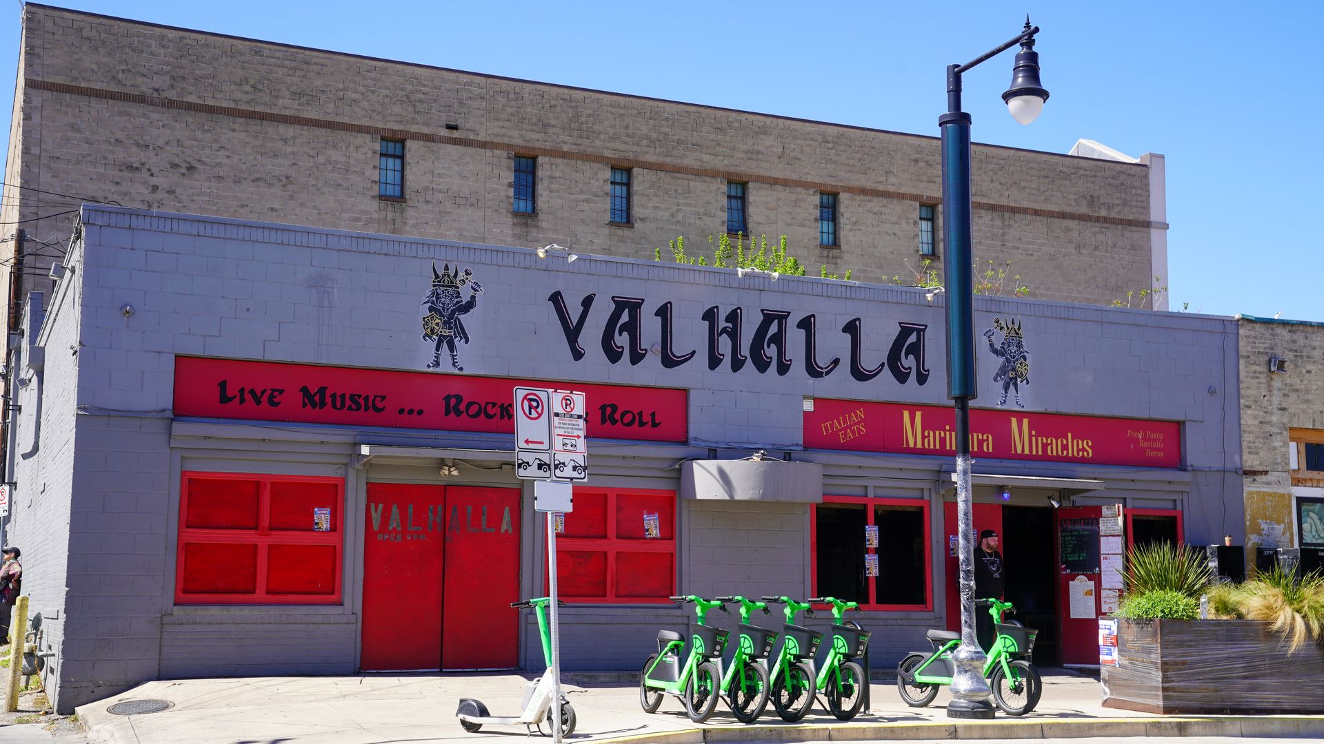 Sunlit street view of a storefront labeled "VALHALLA" with Viking logos, red doors, a red banner, and a row of green bike-share bikes; a blue streetlamp stands nearby under a clear blue sky.