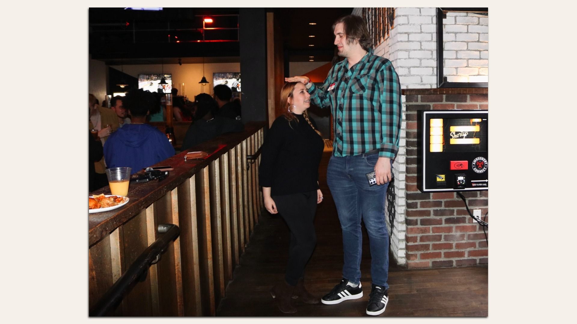 A tall man in a green and black checkered shirt measures a shorter woman’s height by placing his hand above her head in a dimly lit bar with brick walls.