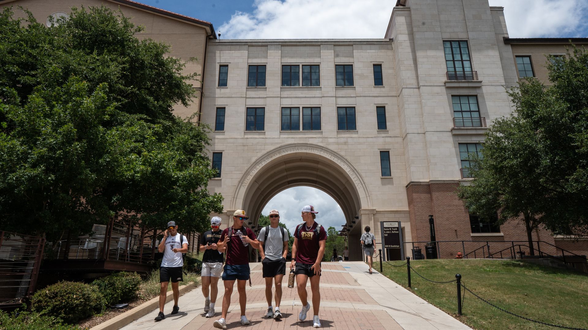 Stone university building with a tall tower and large archway under a blue sky; trees frame the scene as students walk through the arch toward the camera.