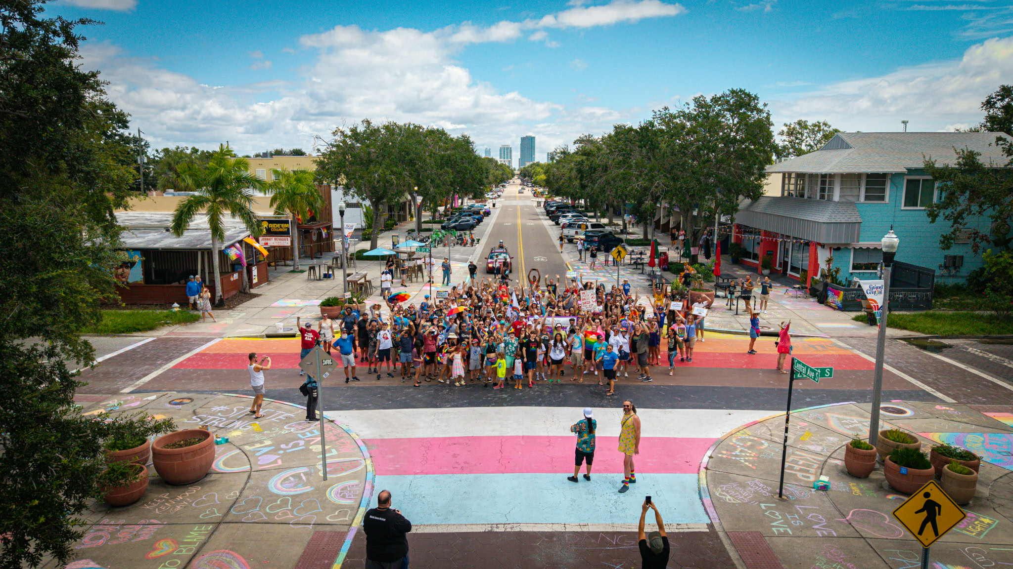 A large group of people gather on a rainbow-painted intersection with colorful chalk art and signs supporting LGBTQ+ rights.
