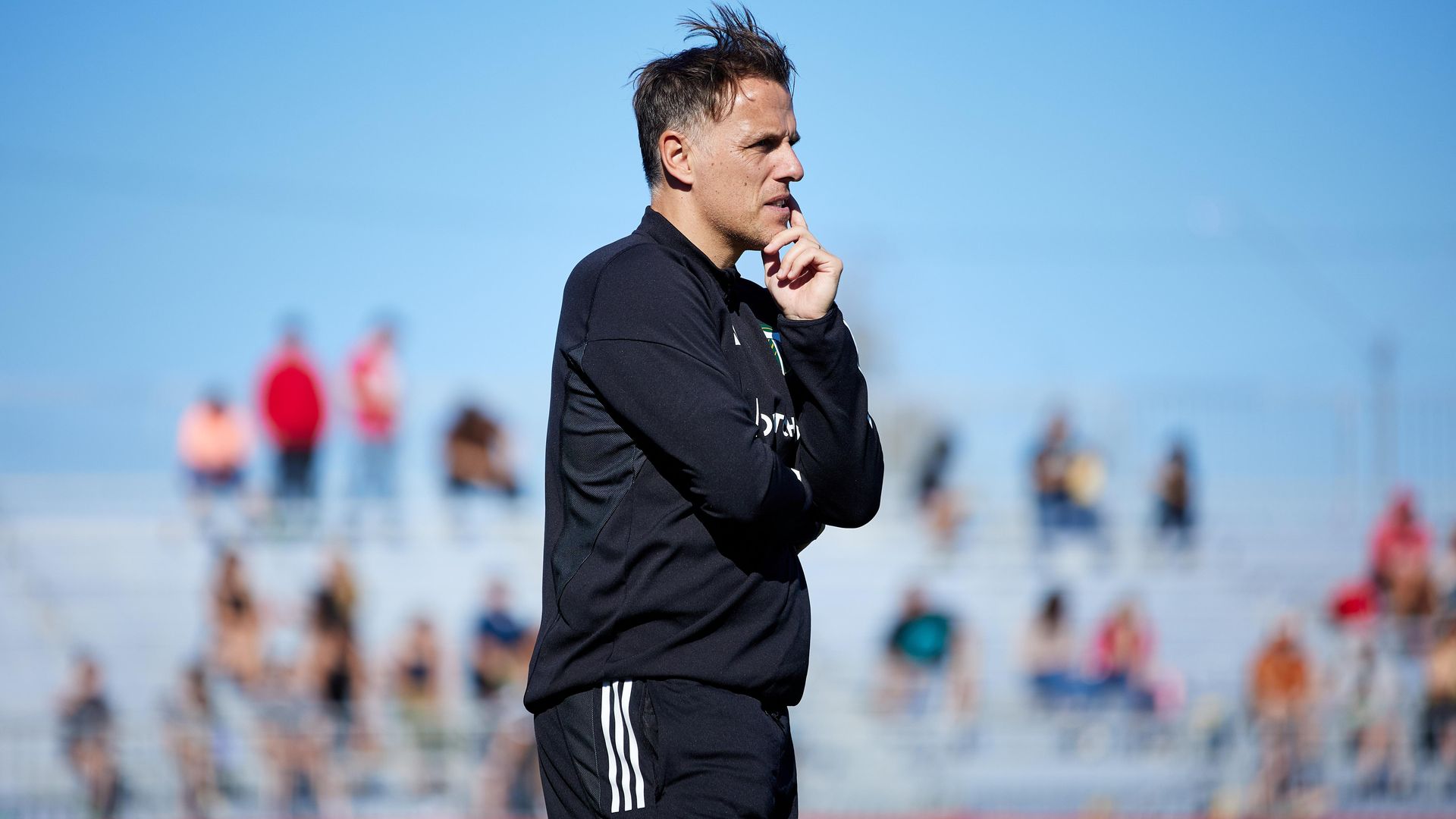 A wite man in sportswear with his chin in his hands looks on at a soccer stadium in Arizona with blurry fans on the  bleachers behind him.