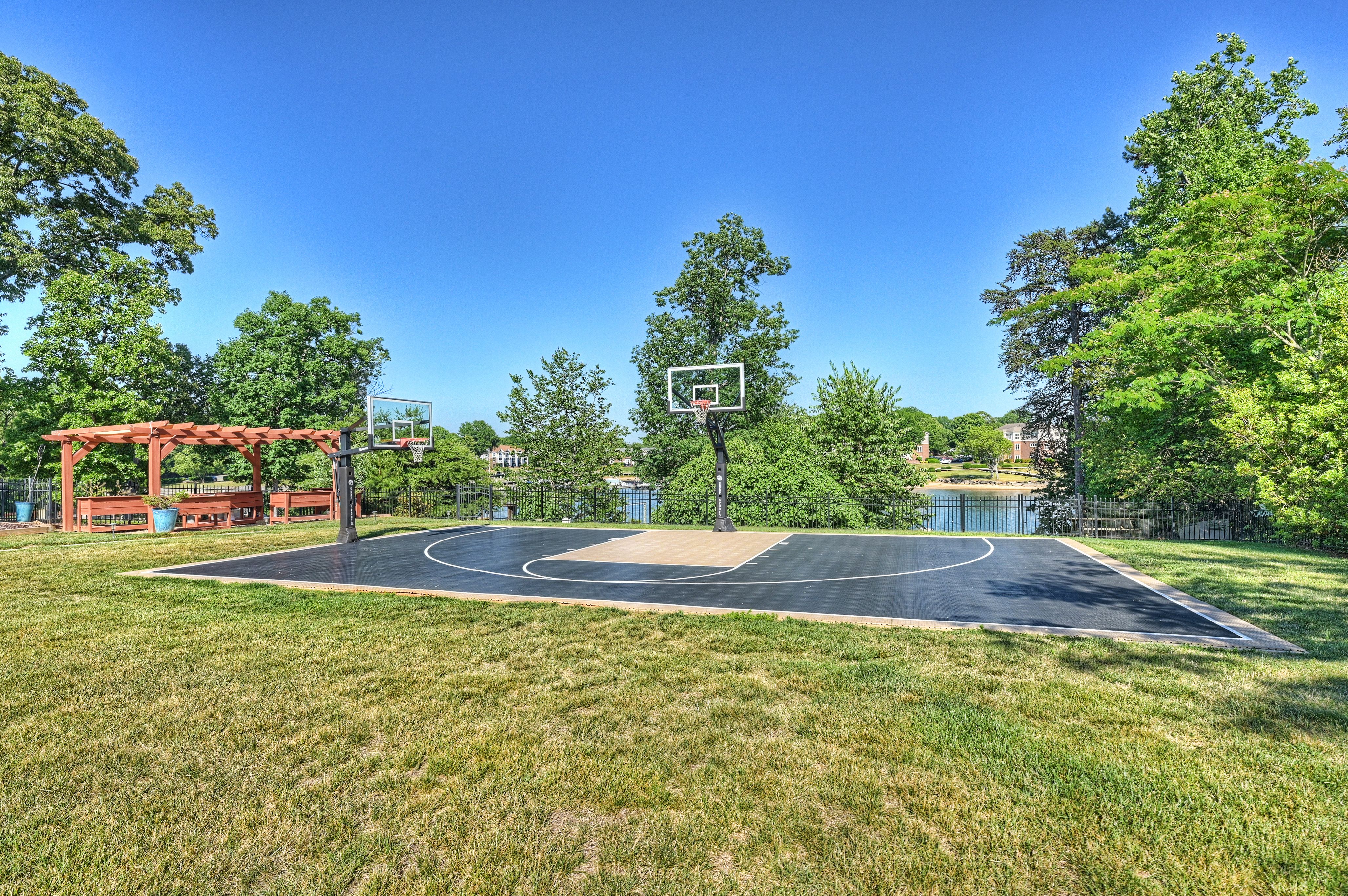 Outdoor basketball court with black asphalt, white lines, and a hoop with backboard. Surrounded by green trees under a bright blue sky; a red wooden pergola sits to the left, with a fence by a lake.