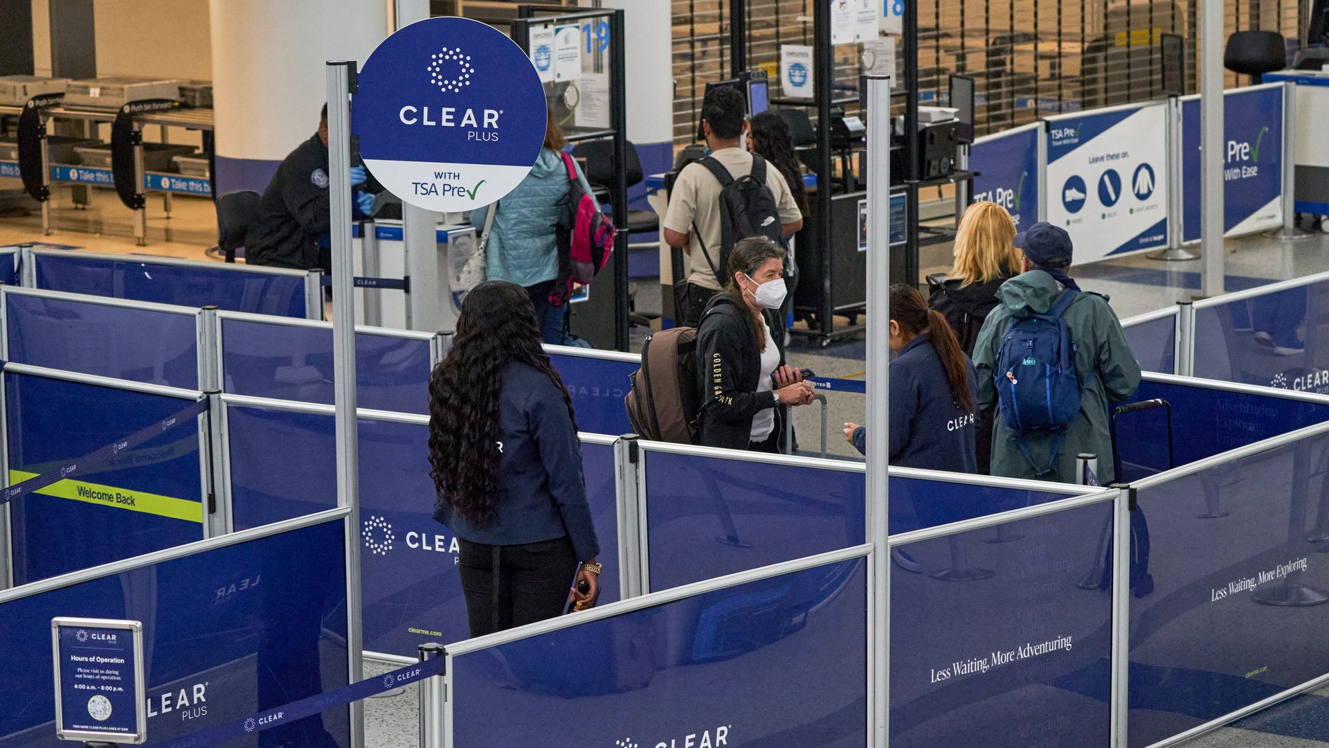 Airport security line for CLEAR Plus with TSA PreCheck, blue barriers, passengers and staff wearing masks, some carrying backpacks.