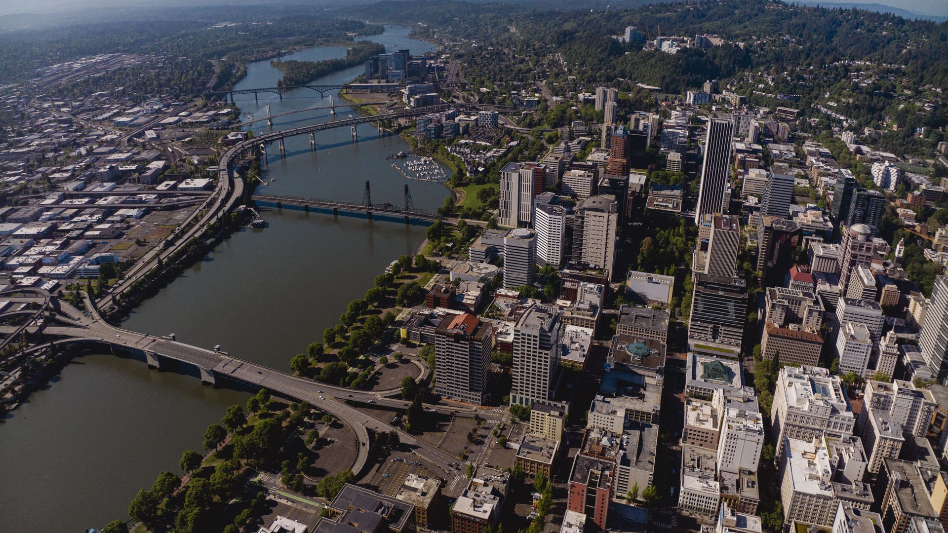 An aerial cityscape of a river through downtown, with bridges, tall modern buildings, roads and greenery, and forested hills in the distance under a clear blue sky.