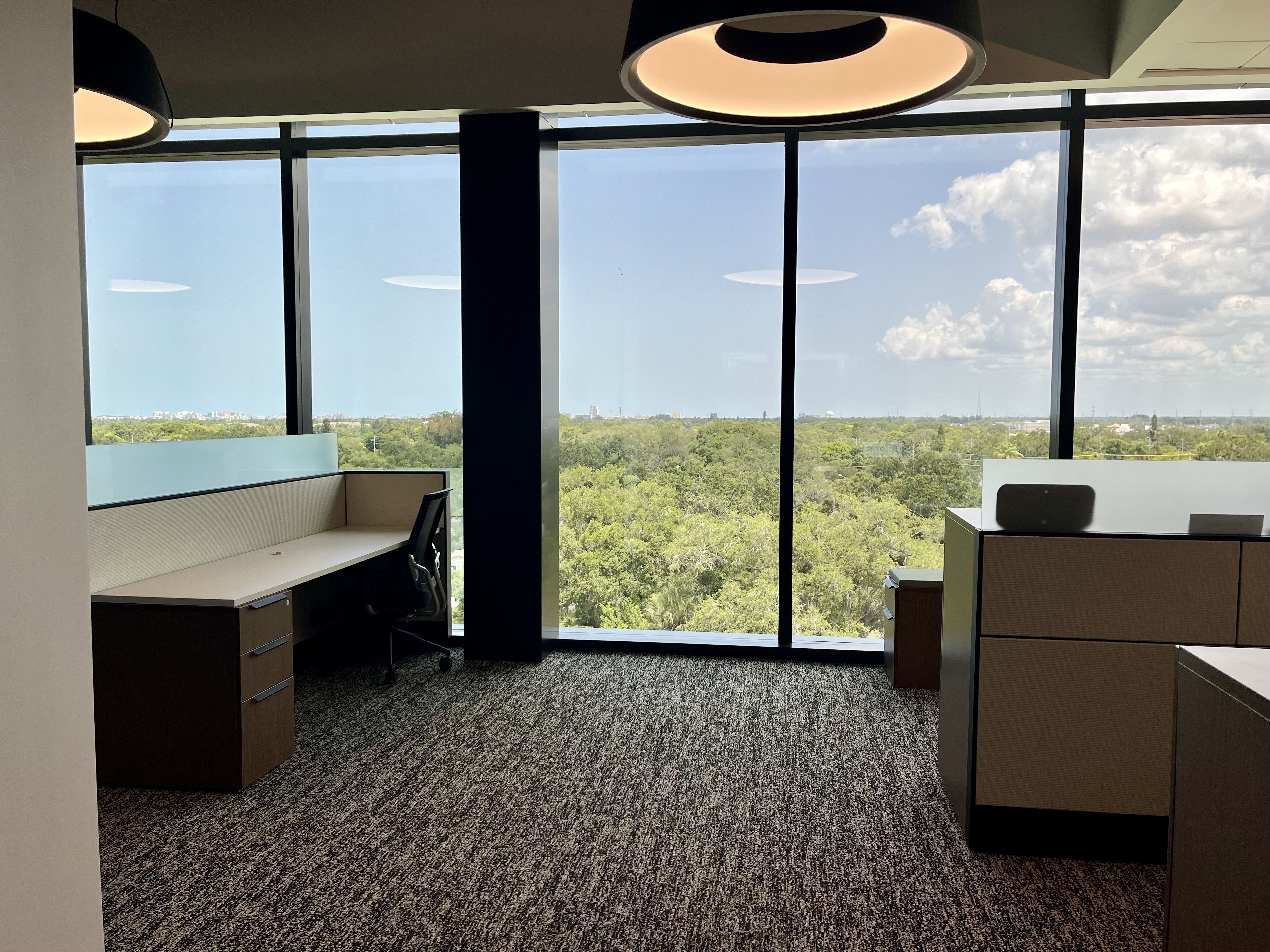 Office interior with large floor-to-ceiling windows showing a green treetop landscape and blue sky; two round pendant lights hang from the ceiling; desks, chairs, and filing cabinets visible.
