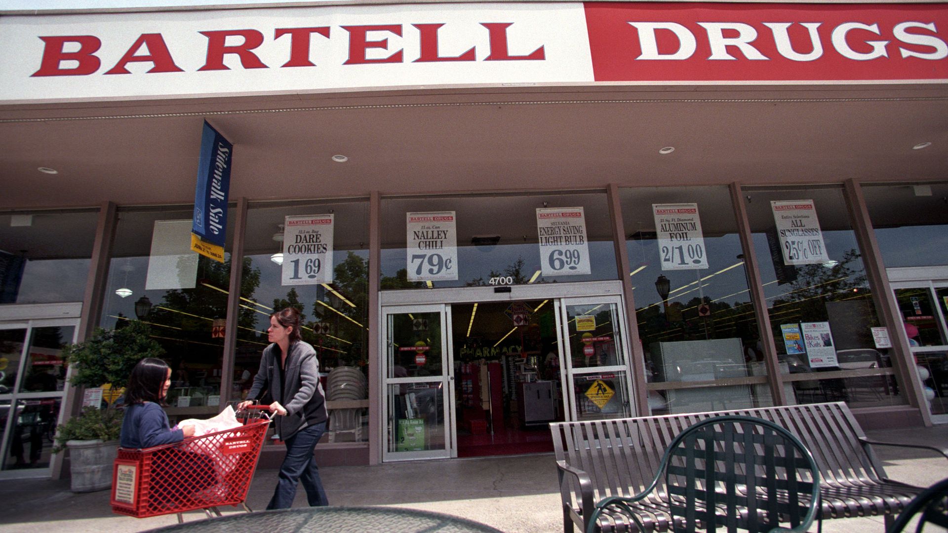 A woman with a child in a shopping cart walks in front of a Bartell Drugs store. 