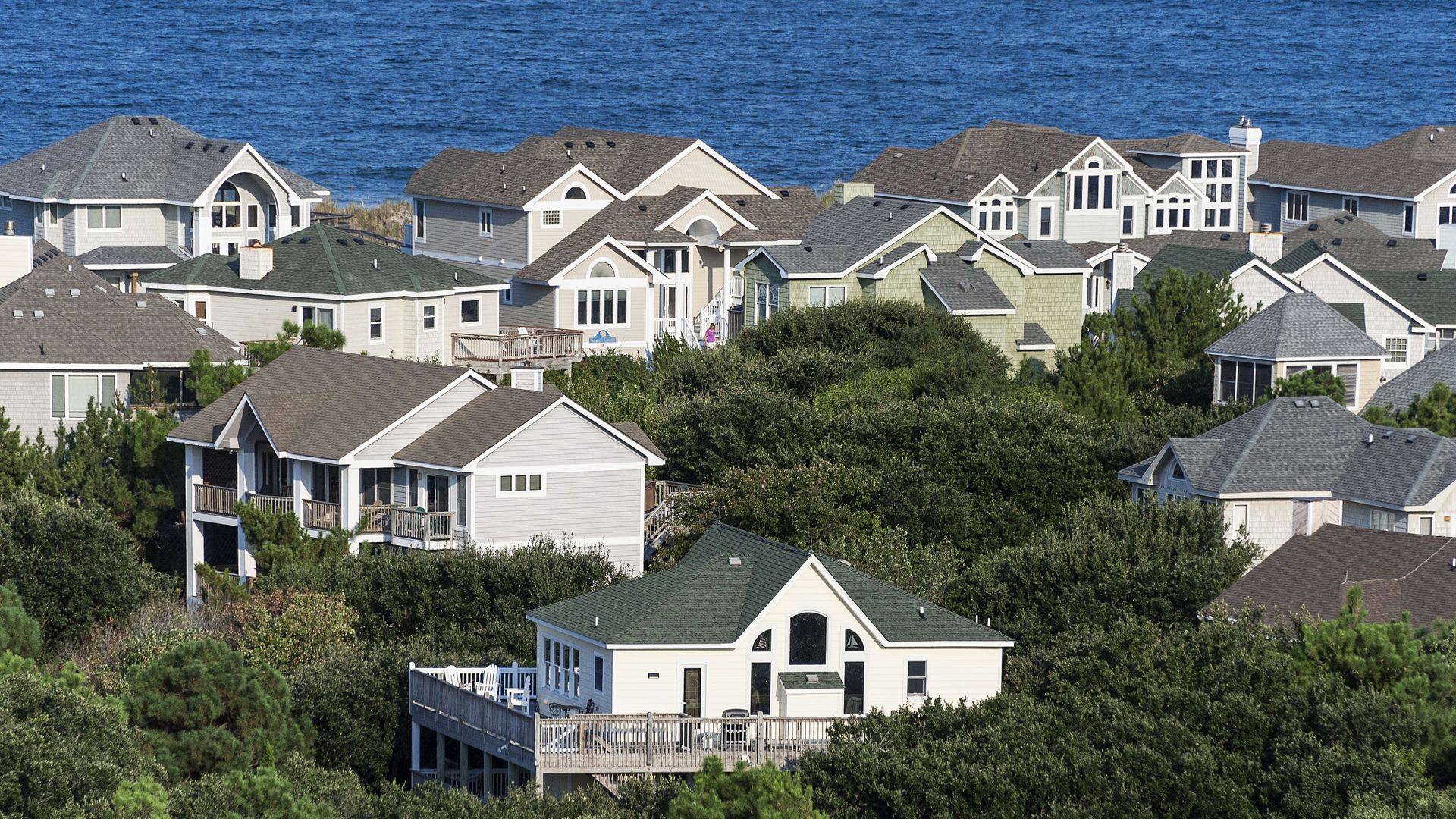 Row of light-colored coastal houses with gabled roofs on a hillside, green trees in the foreground, and a wide blue ocean behind.
