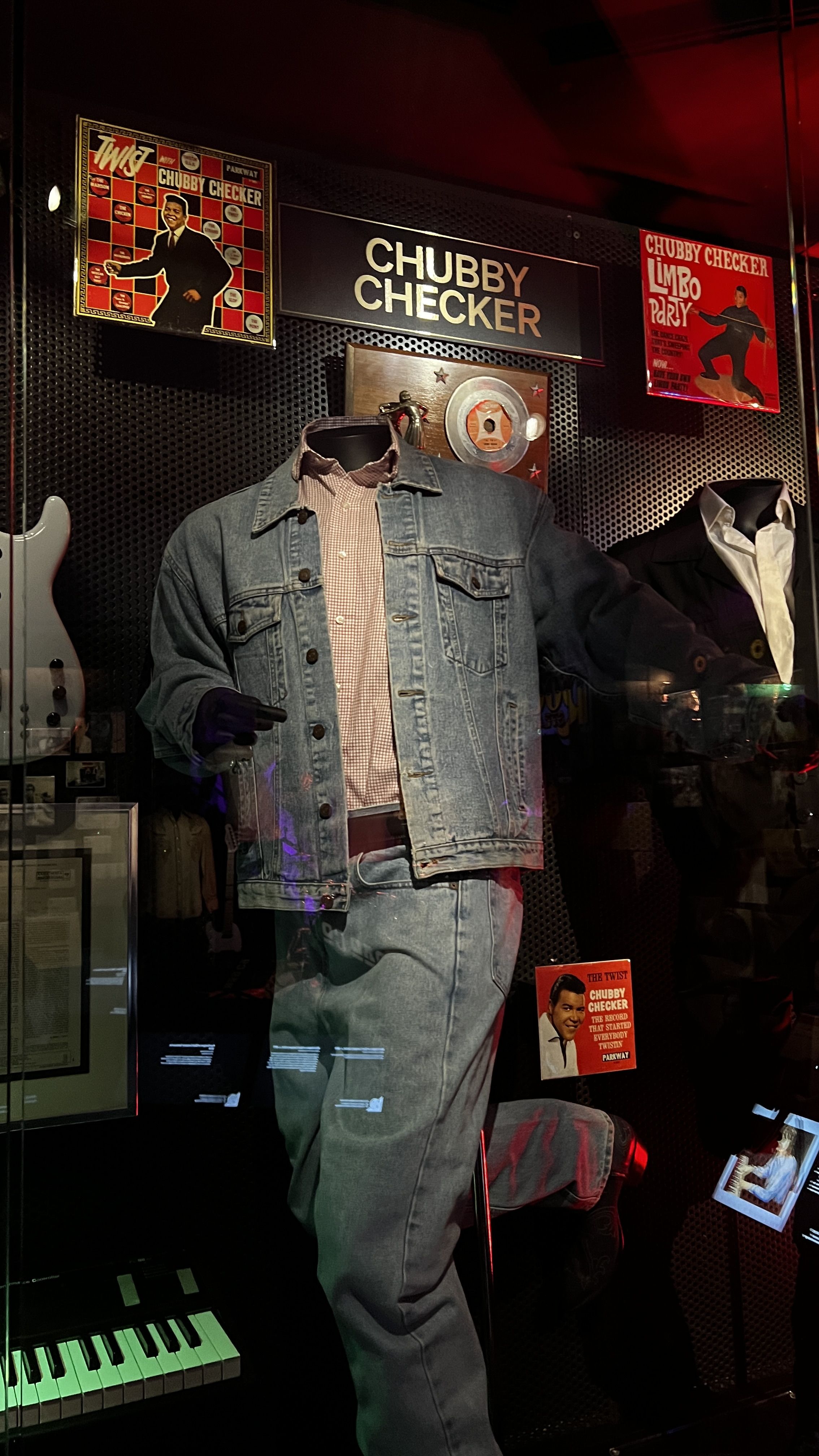 Display case featuring a denim jacket and jeans outfit on a mannequin, with red and black Chubby Checker album covers and a sign with his name in gold letters on a black background.