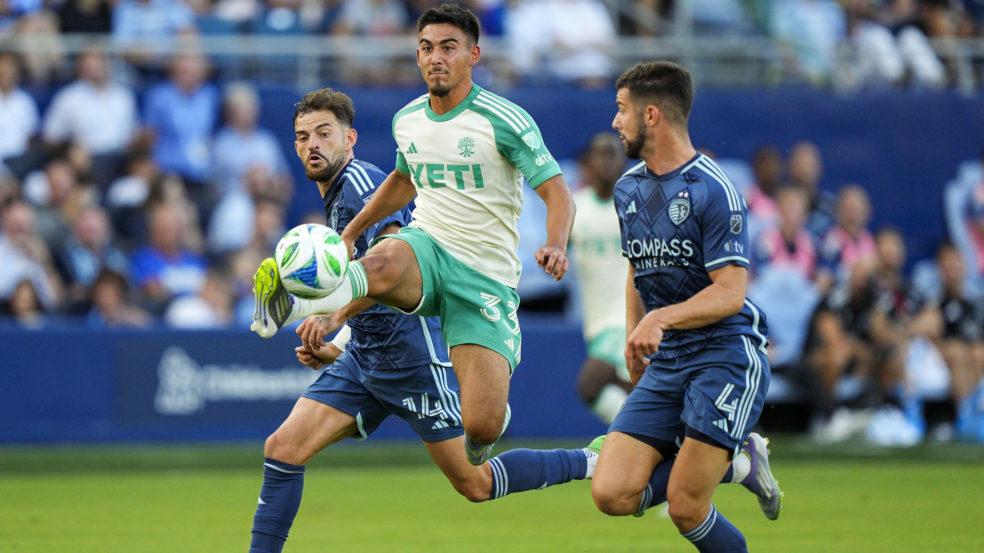 A soccer player in white and green with "YETI" on jersey kicks the ball mid-air, surrounded by two players in dark blue uniforms with "COMPASS" on their jerseys, on a green field with a blurry crowd.