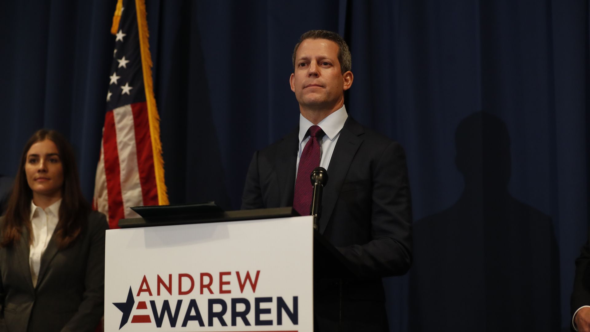 A man stands behind a lectern with a placard that reads "Andrew Warren" in red and blue letters. Beside him stands an American flag.