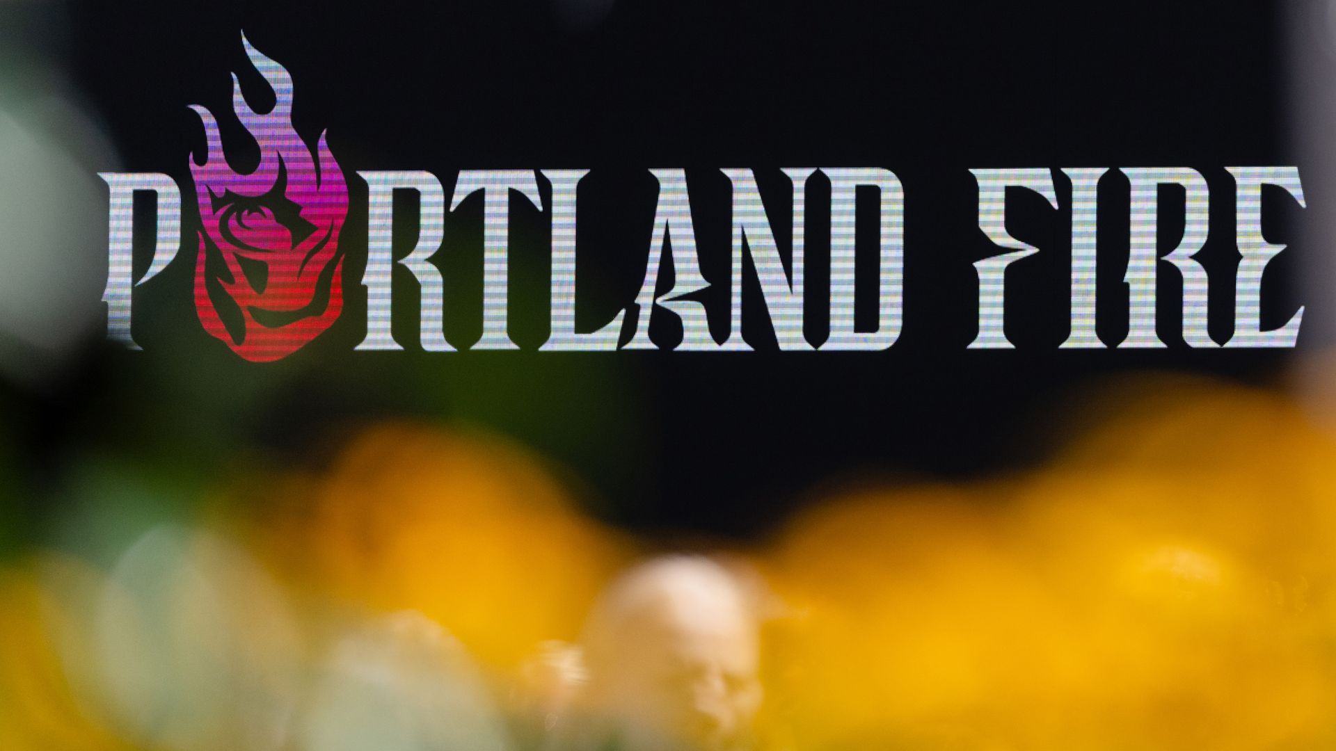 Portland Fire logo with stylized red and purple flame replacing the letter O, against a dark background with blurred yellow and green colors in the foreground.