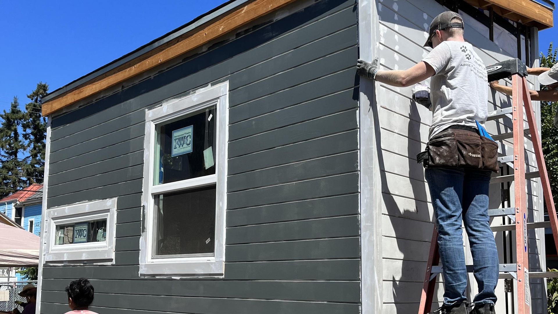 A person on a ladder works on a tiny house painted grey.