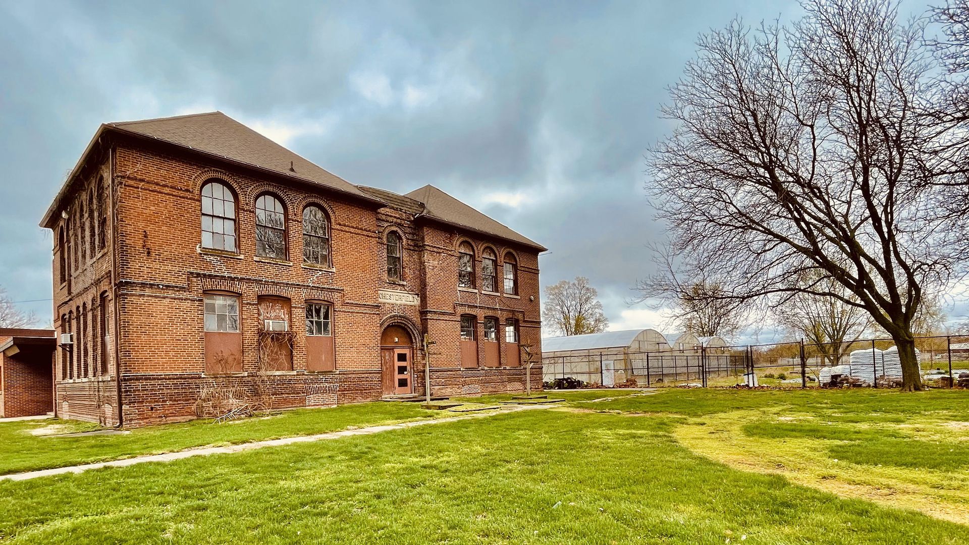 Old two-story red brick building with boarded windows and a sign that reads Chesterfell, surrounded by green grass and leafless trees under a cloudy sky.