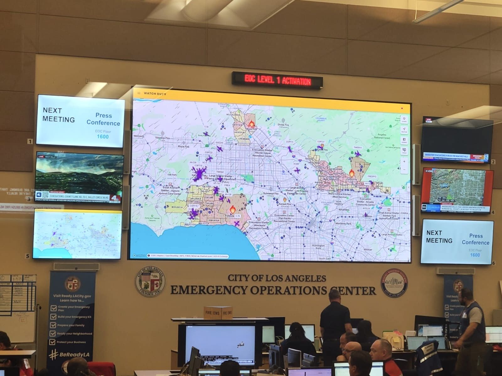 City of Los Angeles Emergency Operations Center with large screens showing wildfire maps and live news footage, staff working at computer stations, and signs for next press conference at 1600 hours.