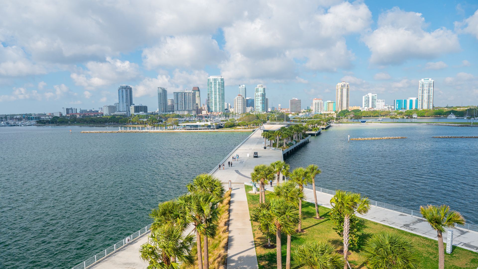 A waterfront cityscape with a long pedestrian pier lined with palm trees, where people stroll toward the skyline of modern high-rise buildings across calm water, under a blue sky with white clouds.