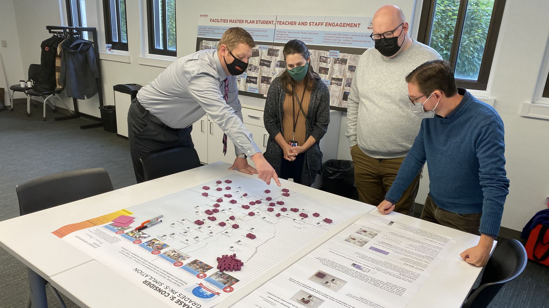 A group of people gathers around a large map on a table, listening to an architect describe it