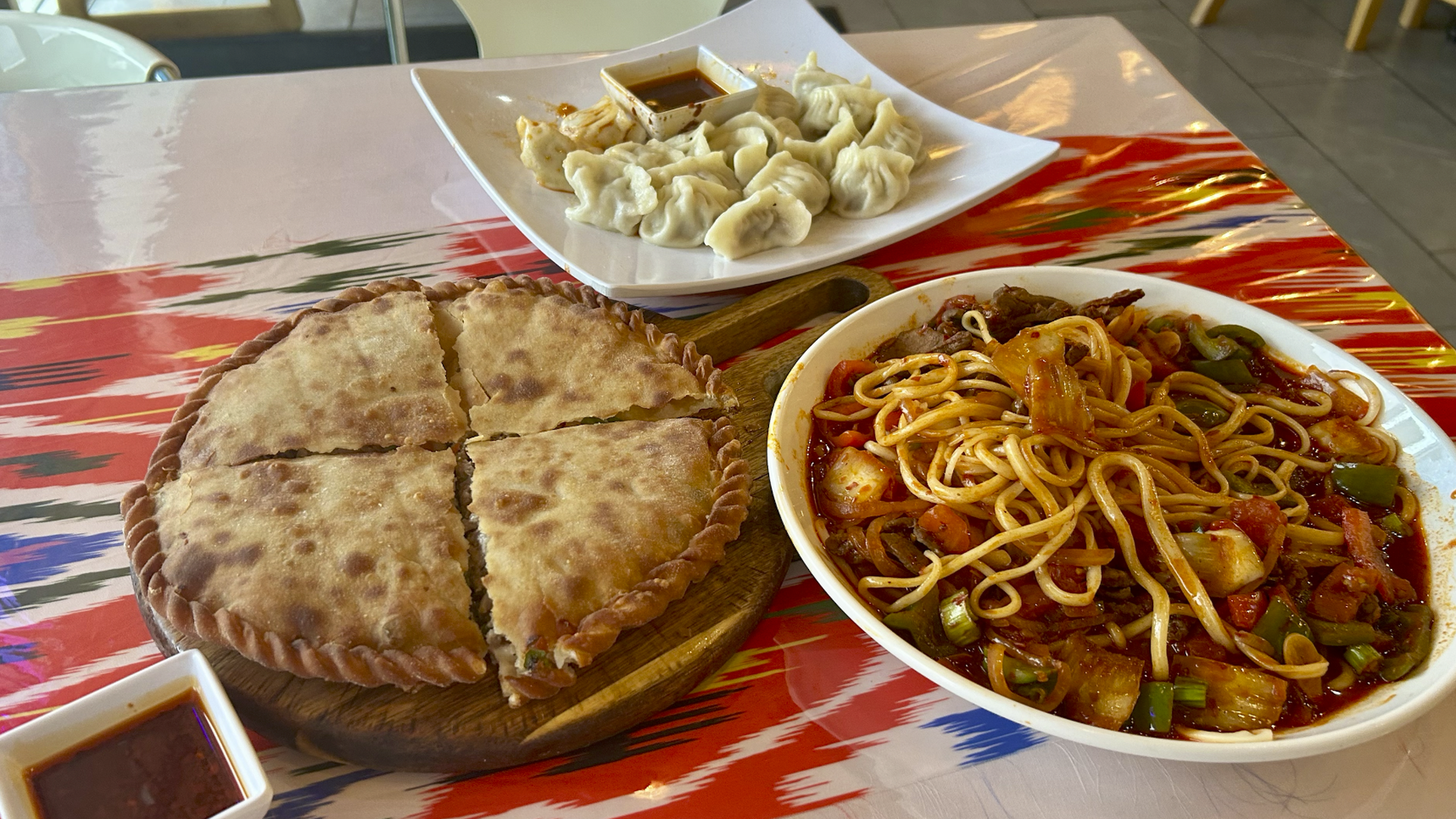 A table with three dishes: a round, sliced flatbread on a wooden board, a white bowl of noodles with vegetables in red sauce, and a plate of steamed dumplings with dipping sauce.
