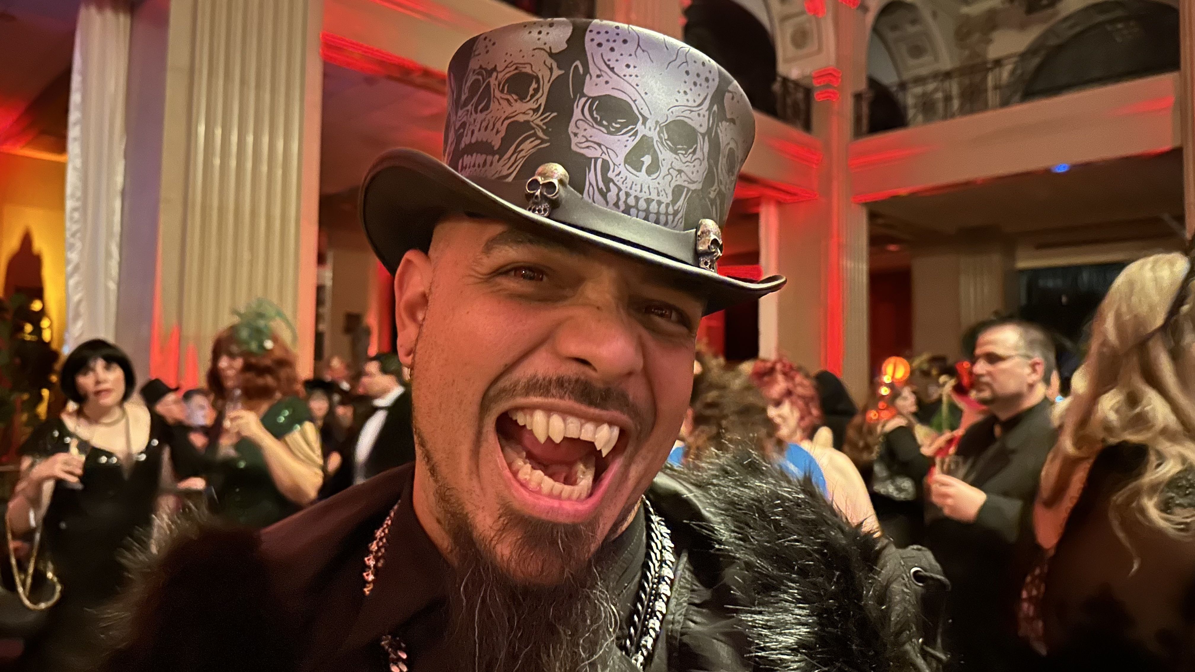 Man at a party wearing a black top hat with skull designs and fangs, smiling widely. Background shows people in costumes in a warmly lit room with red accents and columns.
