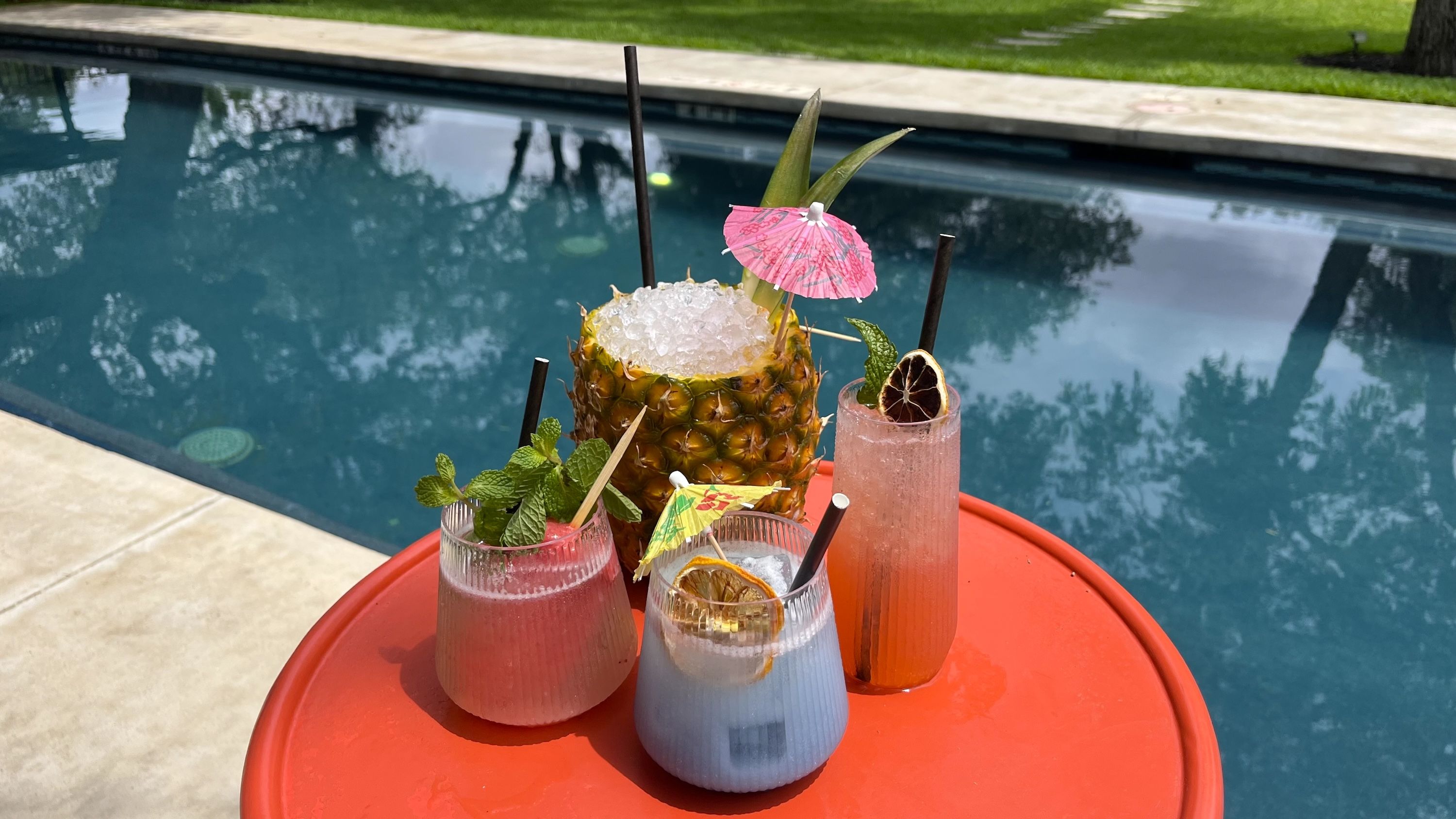 Poolside scene with a pineapple cup of crushed ice and pink cocktails on a bright red table, mint garnish and a pink umbrella; blue pool and green lawn reflected in the water.