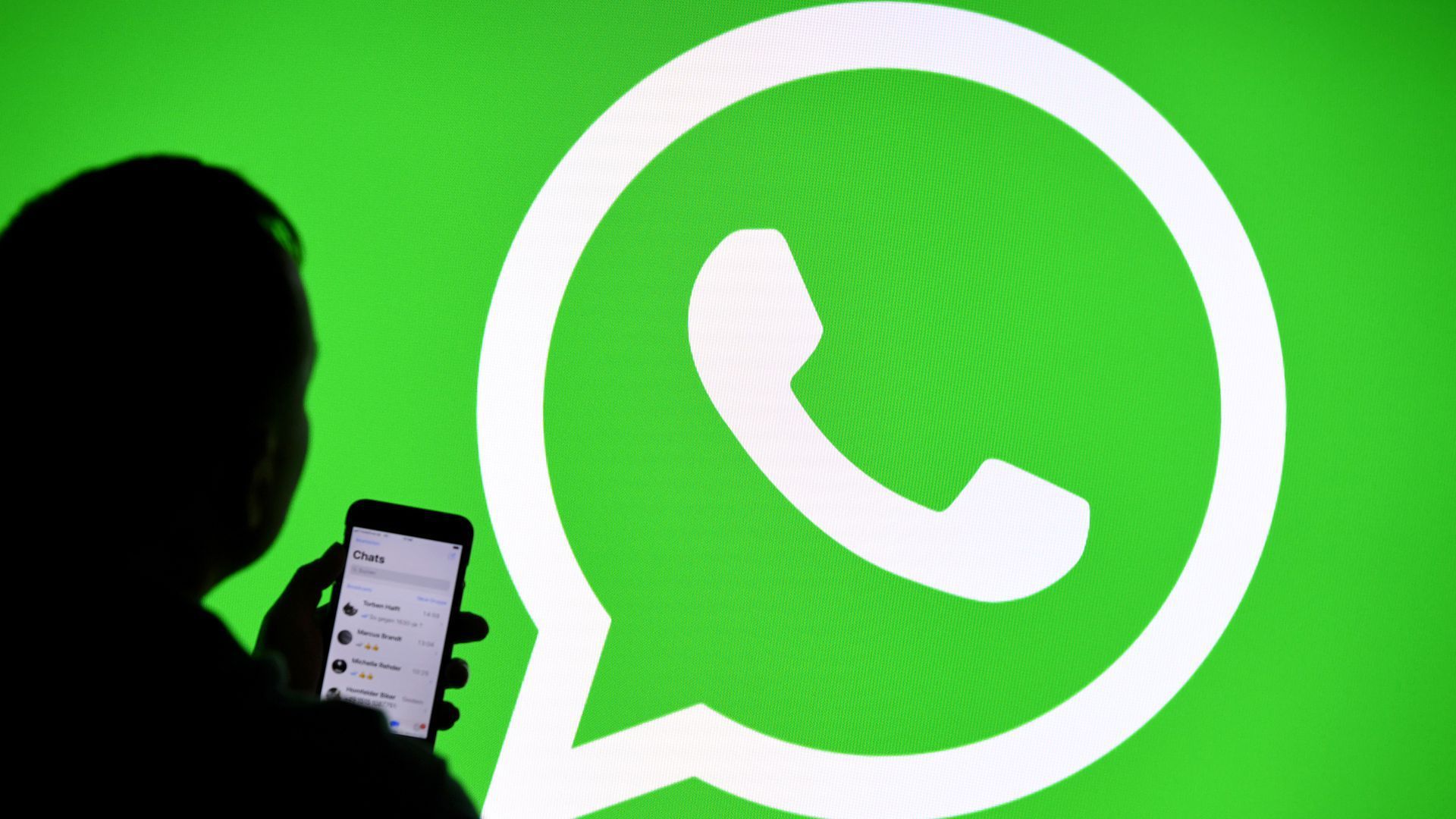 A photo illustration of the silhouette of a person holding a smartphone against a green WhatsApp logo as backdrop