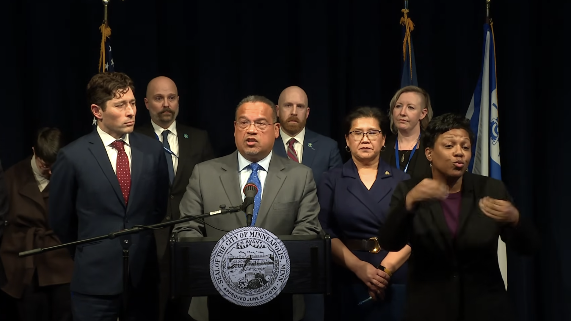 Keith Ellison speaks with Mayors Jacob Frey and Kaohly Her and other people surrounding him. 
