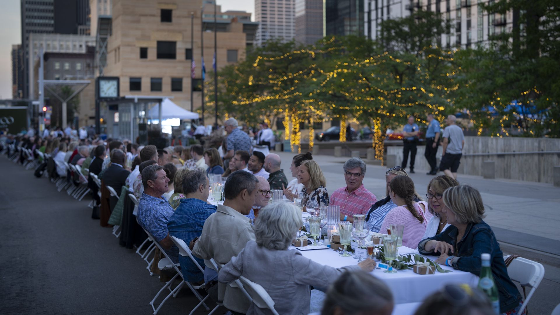 Long outdoor city banquet with dozens of people seated along a white-clothed table on a street, surrounded by skyscrapers, trees with string lights, and a twilight sky.