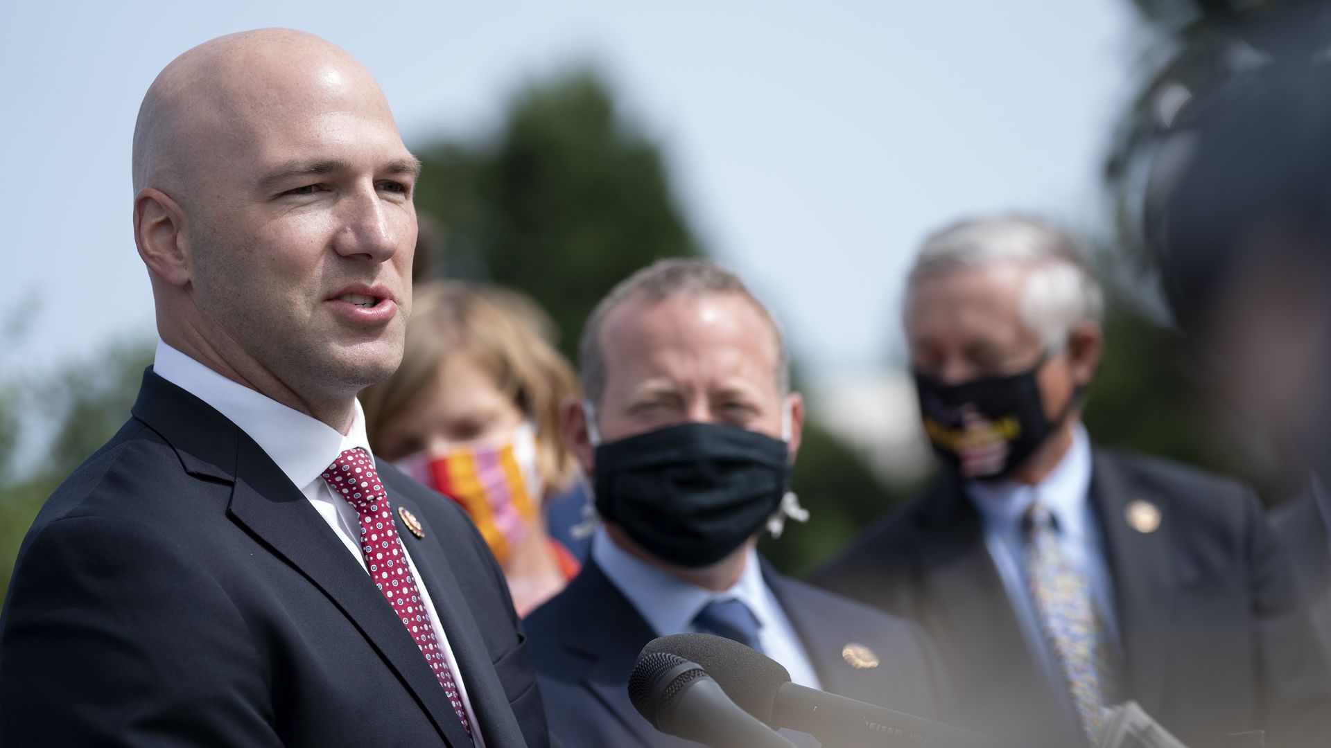 Gonzalez, wearing a suit, is speaking into a microphone while standing outdoors. Other politicians stand in the background.