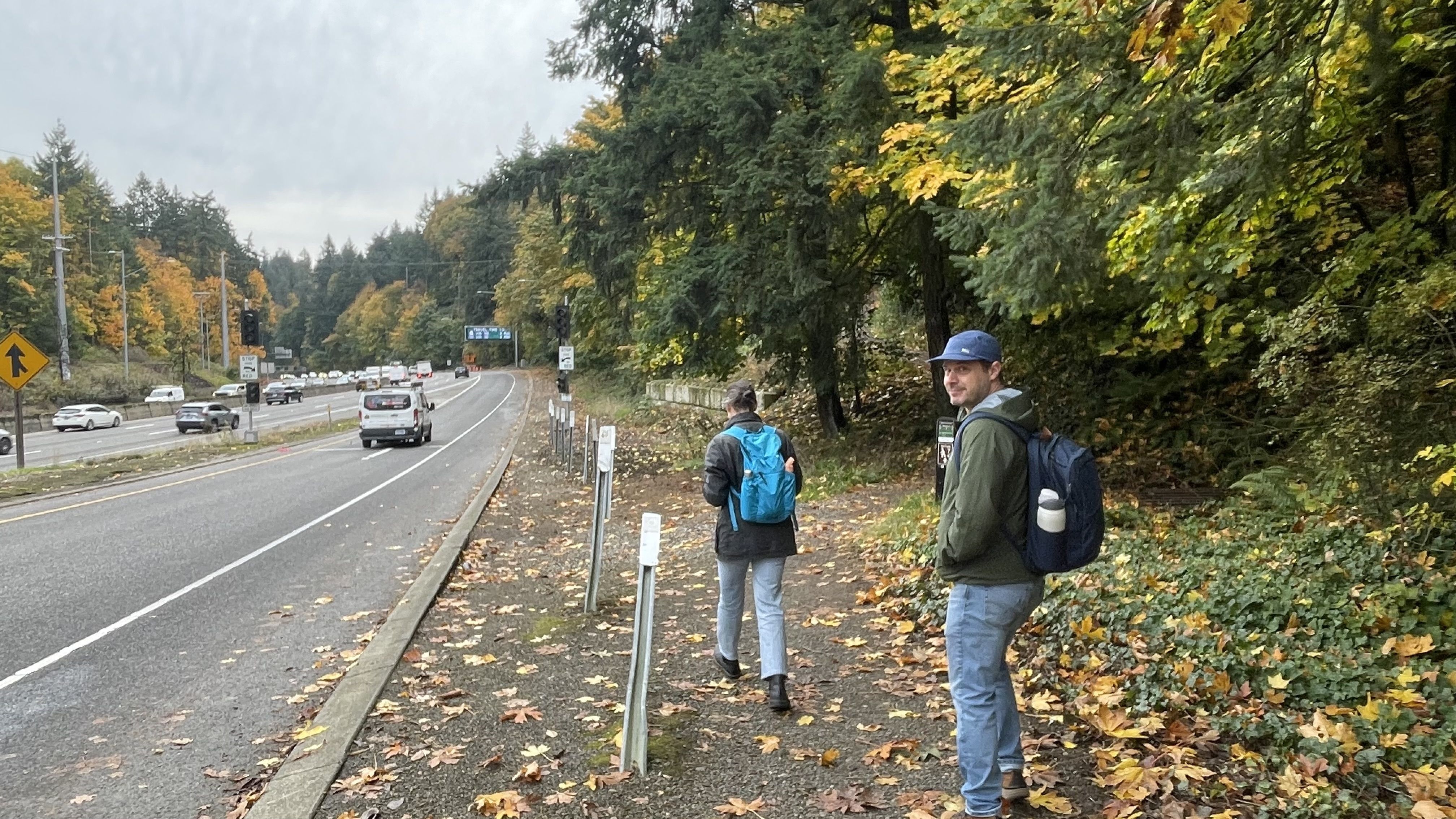 Two people with backpacks walk on a leaf-covered path beside a highway with traffic; green and yellow autumn trees surround the scene under a cloudy sky.