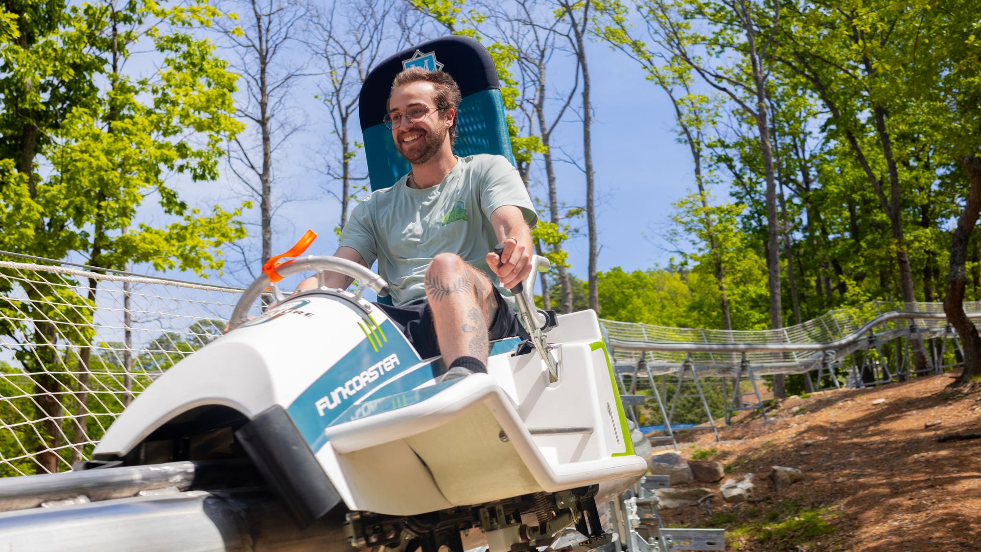 Smiling man with glasses and a beard rides a white and teal vehicle outdoors on a sunny day, with blue sky and green trees behind; a blue headrest and railing are nearby.