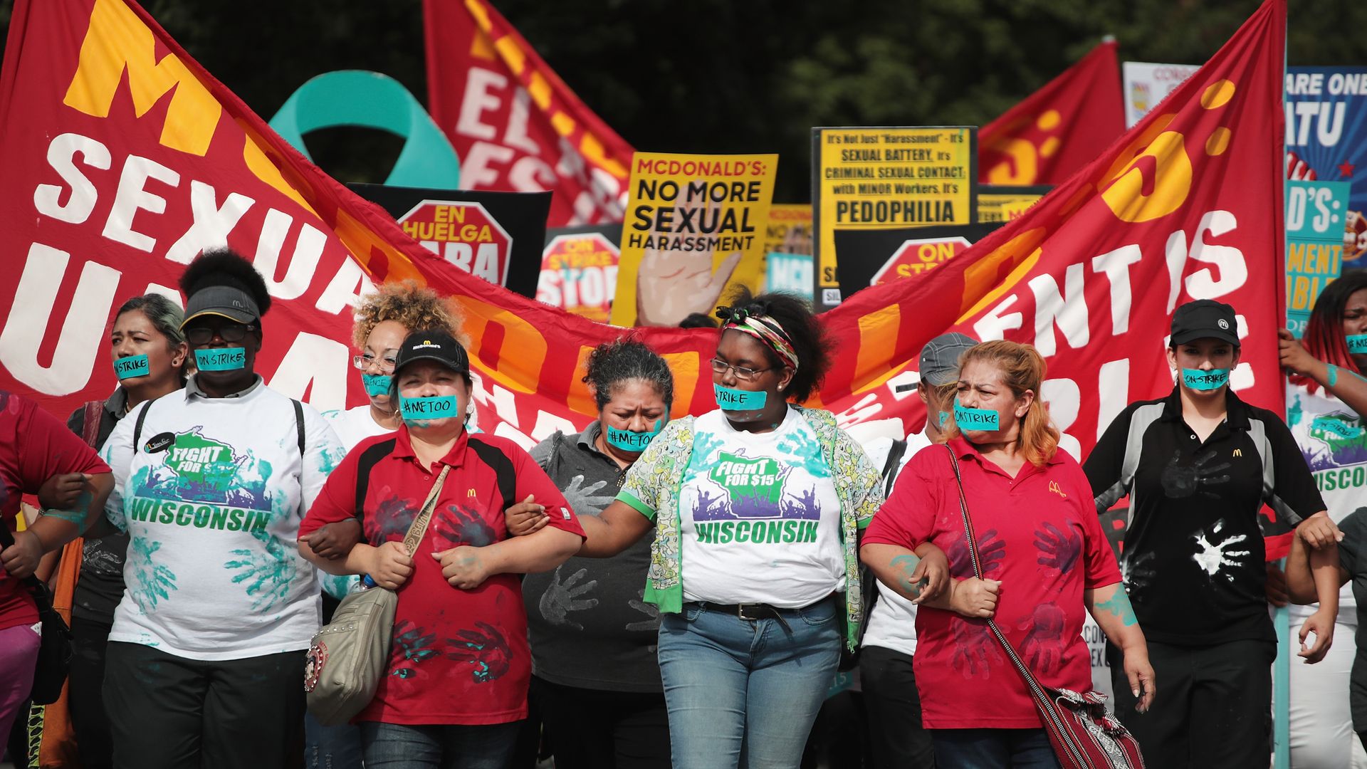In this image, a line of protestors link arms while standing in front of a red banner 