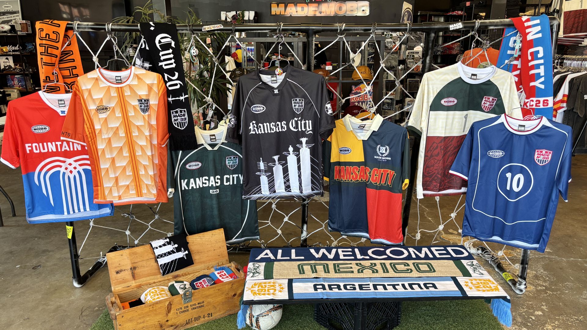 Colorful Kansas City soccer jerseys hang on a metal rack in a shop, with scarves, nets, and crates on the floor; a MADE MOBB sign appears in the background.