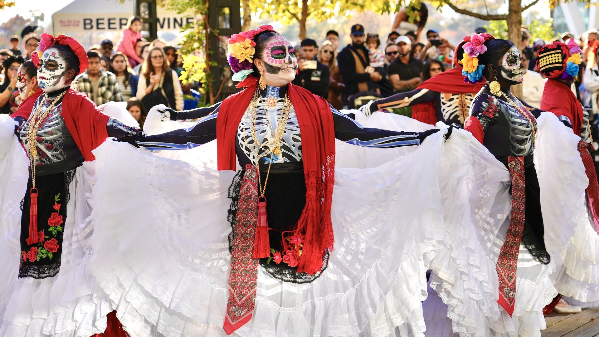 Group of dancers in colorful Day of the Dead skeleton costumes and white skirts, performing outdoors with fall trees and crowd watching in background.