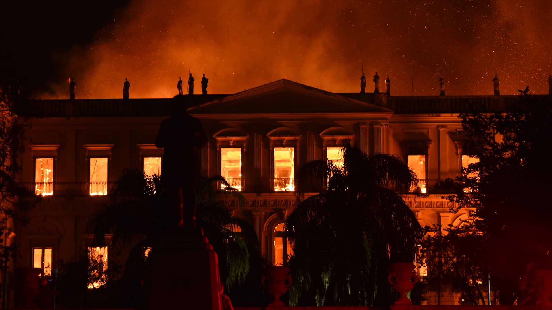 A beautiful museum with sculptures on the top is ablaze. The whole sky looks red and you can see fire through the large windows at the front of the old building. 