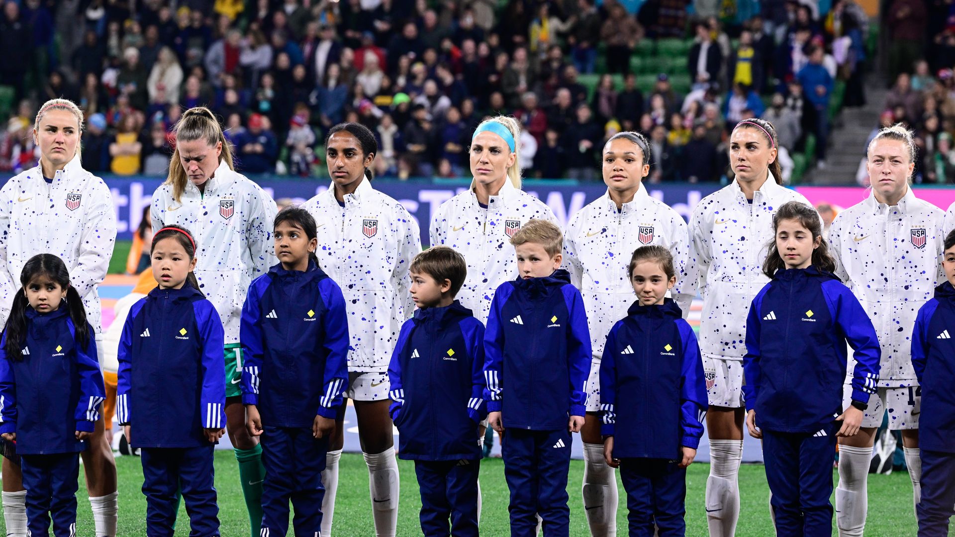 U.S. women's national team players in white warmup jackets stand on the field behind kids in blue track suits at the 2023 FIFA Women's World Cup.