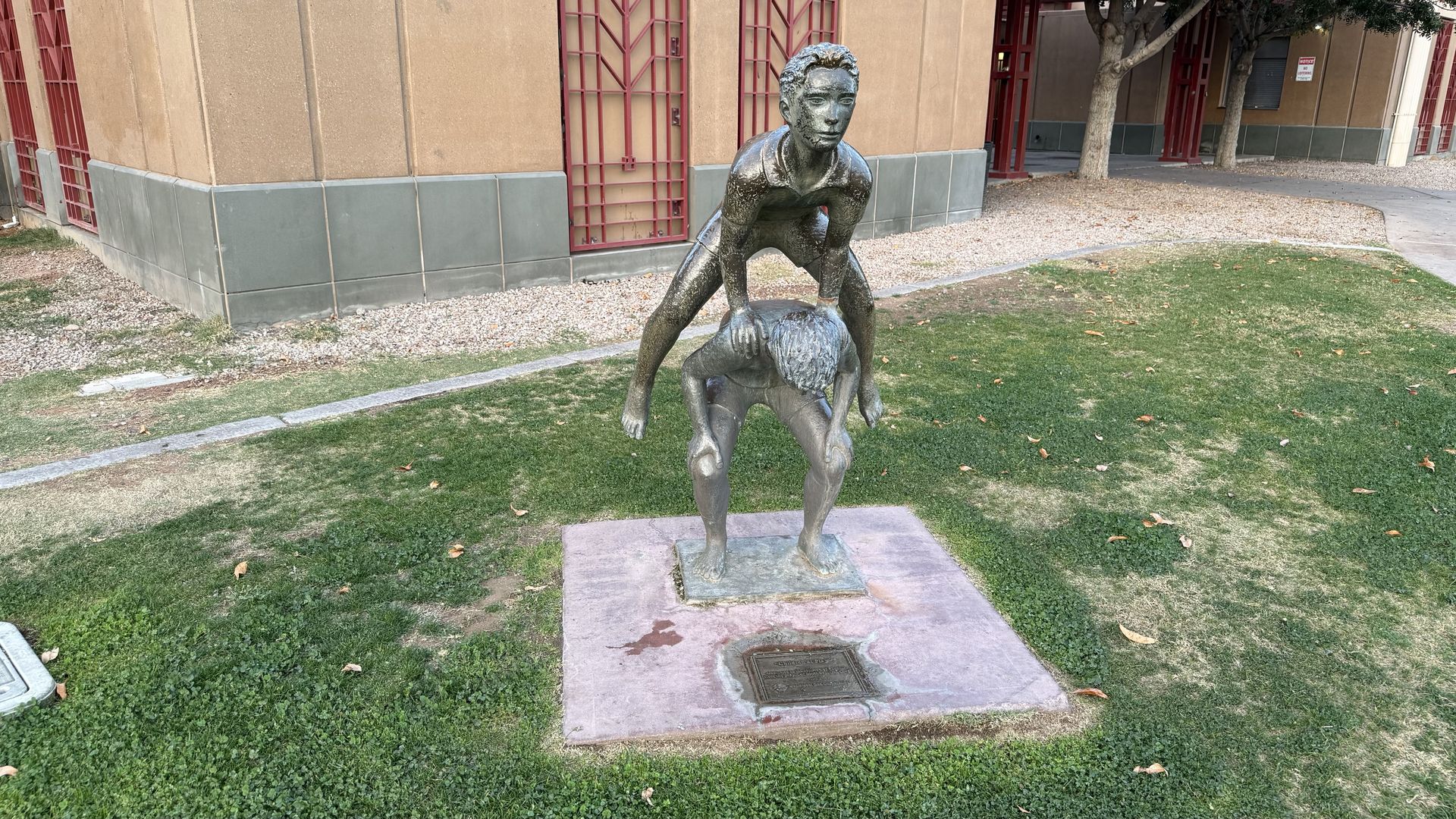 Bronze statue of a child leaping over another child child who's leaning downward, on a square pedestal amid a grassy area with a beige building, red window bars, and trees in the background.