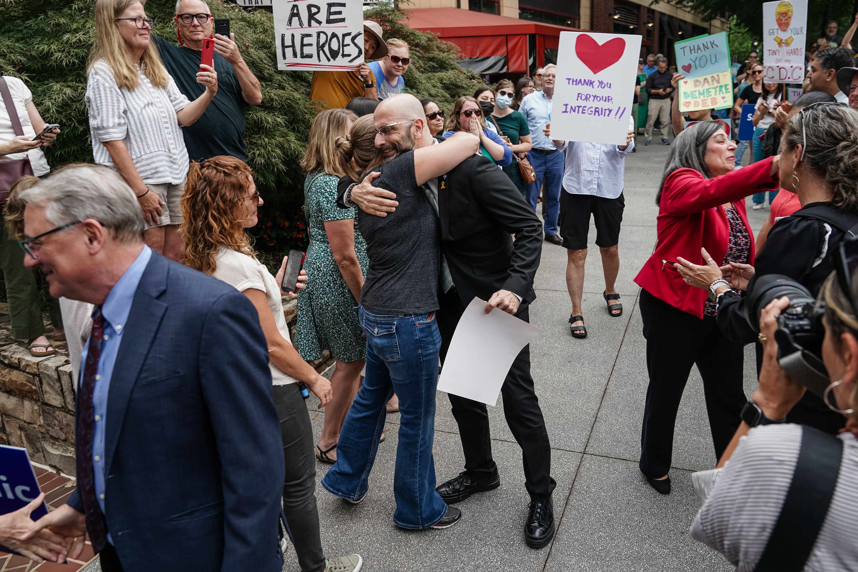 Crowd at outdoor event with people hugging, holding signs reading "Thank you for your integrity!!", "You are heroes", and others, capturing supportive and appreciative atmosphere.