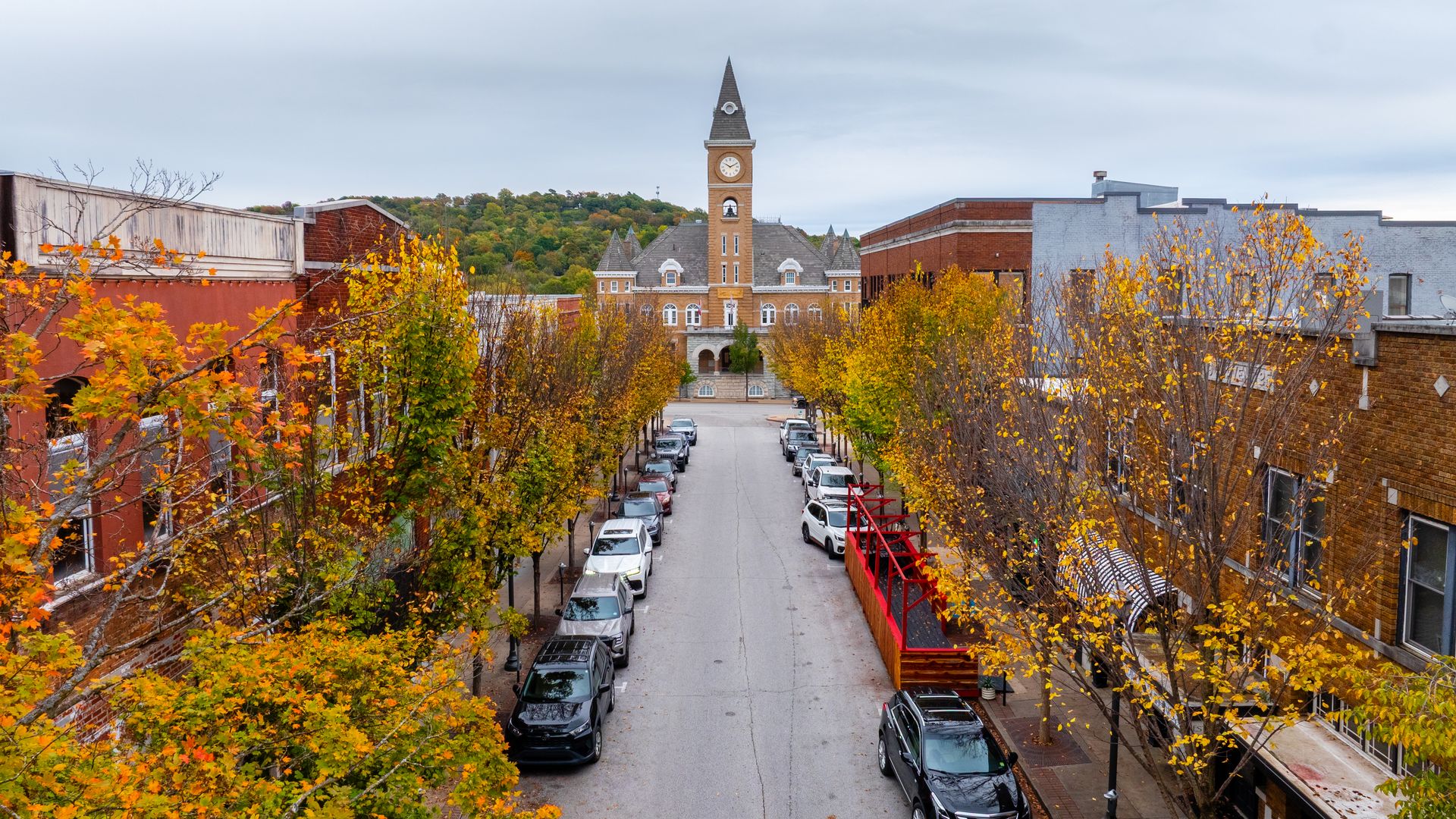 A street view of a small town with a tall clock tower at center, flanked by brick buildings and colorful autumn trees. An overcast sky looms above.
