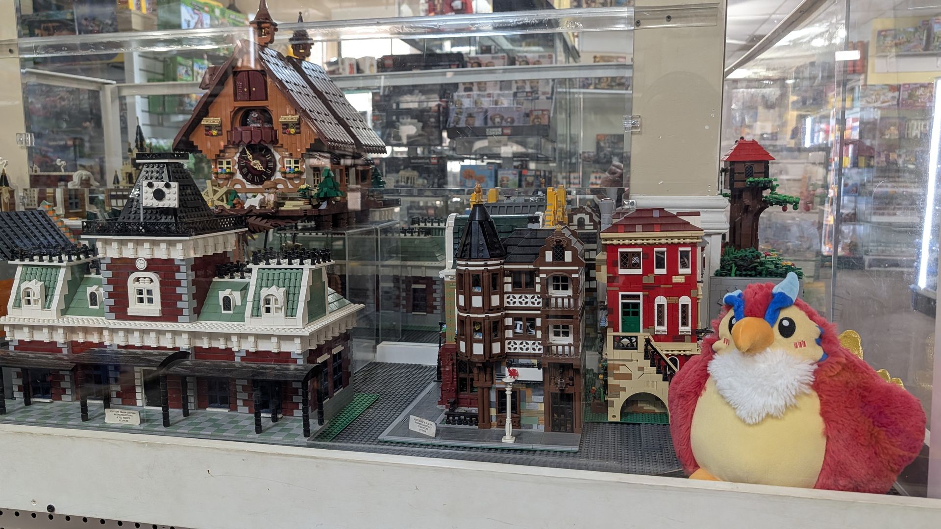 Display case with detailed LEGO buildings and a red, yellow, and blue plush bird toy on the right side, all inside a store filled with more LEGO boxes and toys.
