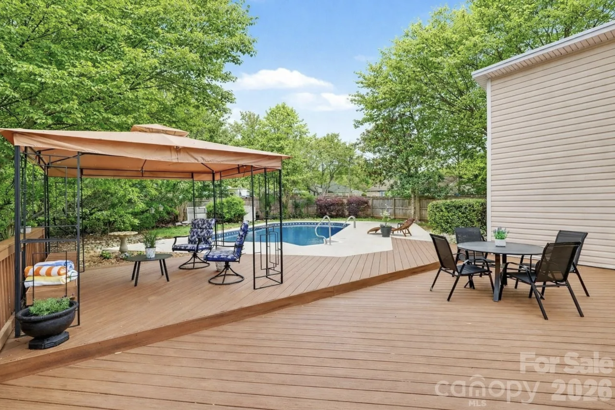 Backyard deck with an orange canopy over a seating area, blue cushions, and potted plants. A curved pool with ladder sits nearby; a round table and chairs are near a beige house.