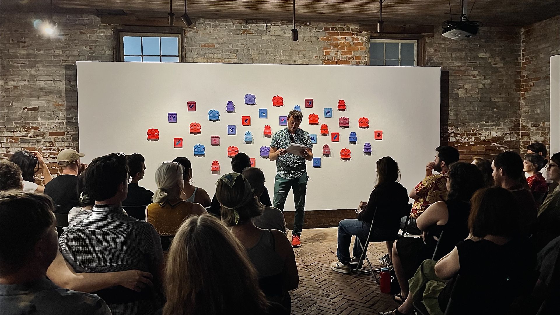 An individual reads a passage from a book to an audience sitting in a room with historic brick walls. A large white gallery wall covered with several rows of adhesive notes is behind the individual reading.