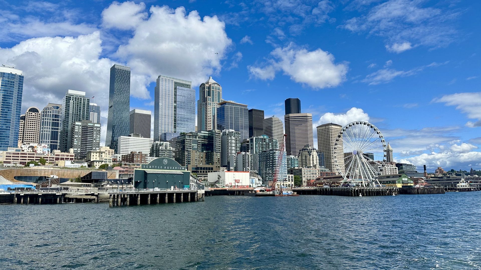 Seattle waterfront with blue water, the Seattle Aquarium building, a large Ferris wheel, and a city skyline of tall buildings under a bright blue sky with white clouds.