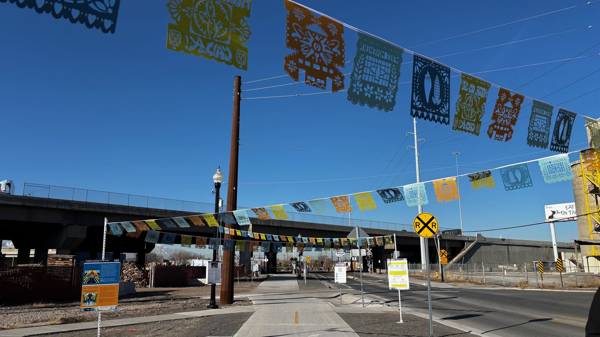 Colorful papel picado decorations hanging across a street and sidewalk near railroad tracks and overpass on a clear sunny day.